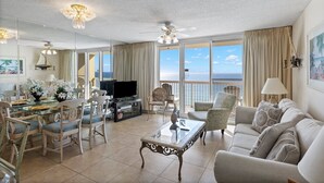 A spacious living room with a glass dining table, comfortable seating, a flat-screen TV, and a balcony with a view of the sea. - Pelican Beach 1202 1 Bedroom Condo by Pelican Beach Management (Destin)
