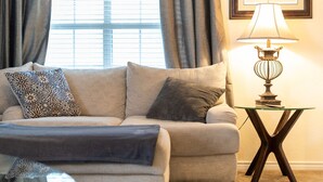 A living room with a beige sofa, patterned pillows, a glass coffee table, and a table lamp.