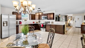 A modern kitchen with stainless steel appliances, dark wood cabinets, a glass dining table, and a chandelier.