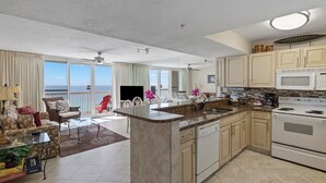 A spacious living area with a large window overlooking the ocean, a kitchen with a central island, and a dining area with a red chair. - Pelican Beach 1406 1 Bedroom Condo by Pelican Beach Management (Destin)