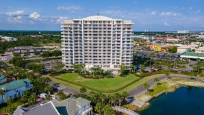 A high-rise hotel with multiple balconies, surrounded by a golf course, parking areas, and a body of water. - Terrace at Pelican Beach 1005 2 Bedroom Condo by Pelican Beach Management (Destin)