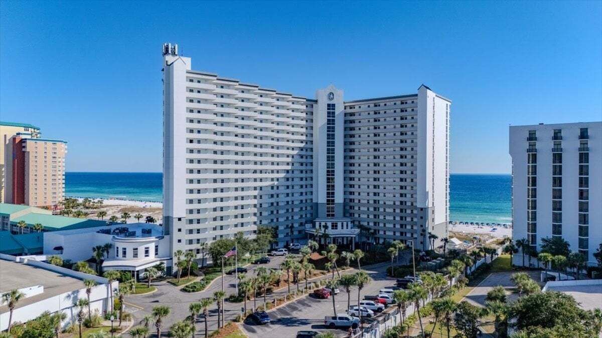 Et højt hotel med flere etager, en parkeringsplads og udsigt til stranden.