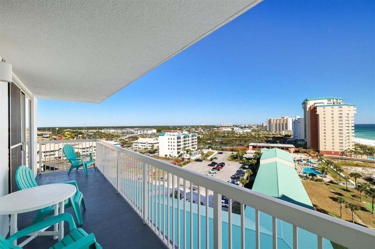 A balcony with turquoise chairs and a table, overlooking a beach and cityscape with hotels and a parking lot.