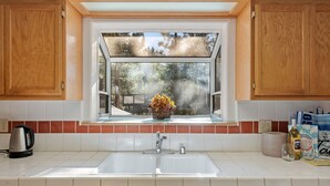 Una cocina con armarios de madera, encimera blanca, ventana con vistas a árboles, un hervidor, una cesta con flores y una bolsa de compras.