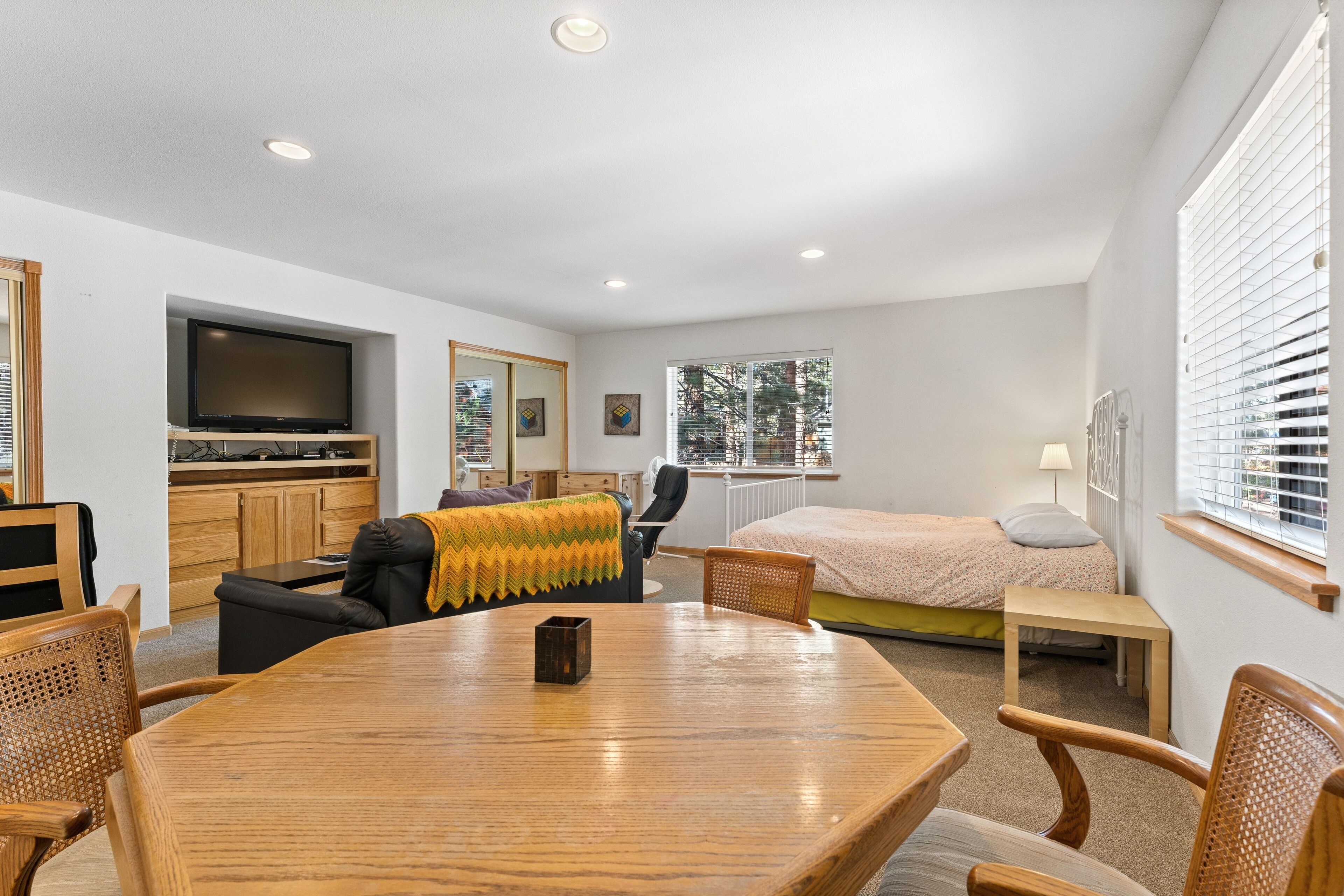 Een moderne, goed verlichte kamer met een houten eettafel, stoelen, een bank, een bed, een televisie en een raam met jaloezieën.