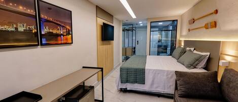 A modern hotel room with a bed, a desk, a small refrigerator, and a TV.
