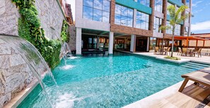 A swimming pool with a waterfall feature, surrounded by a modern building with large windows and outdoor seating. - Gaeta Hotel (Guarapari)