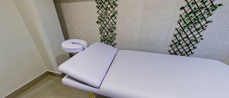 A clinical examination room with a white examination table, a footrest, a wall-mounted plant, and a toilet with a paper roll.