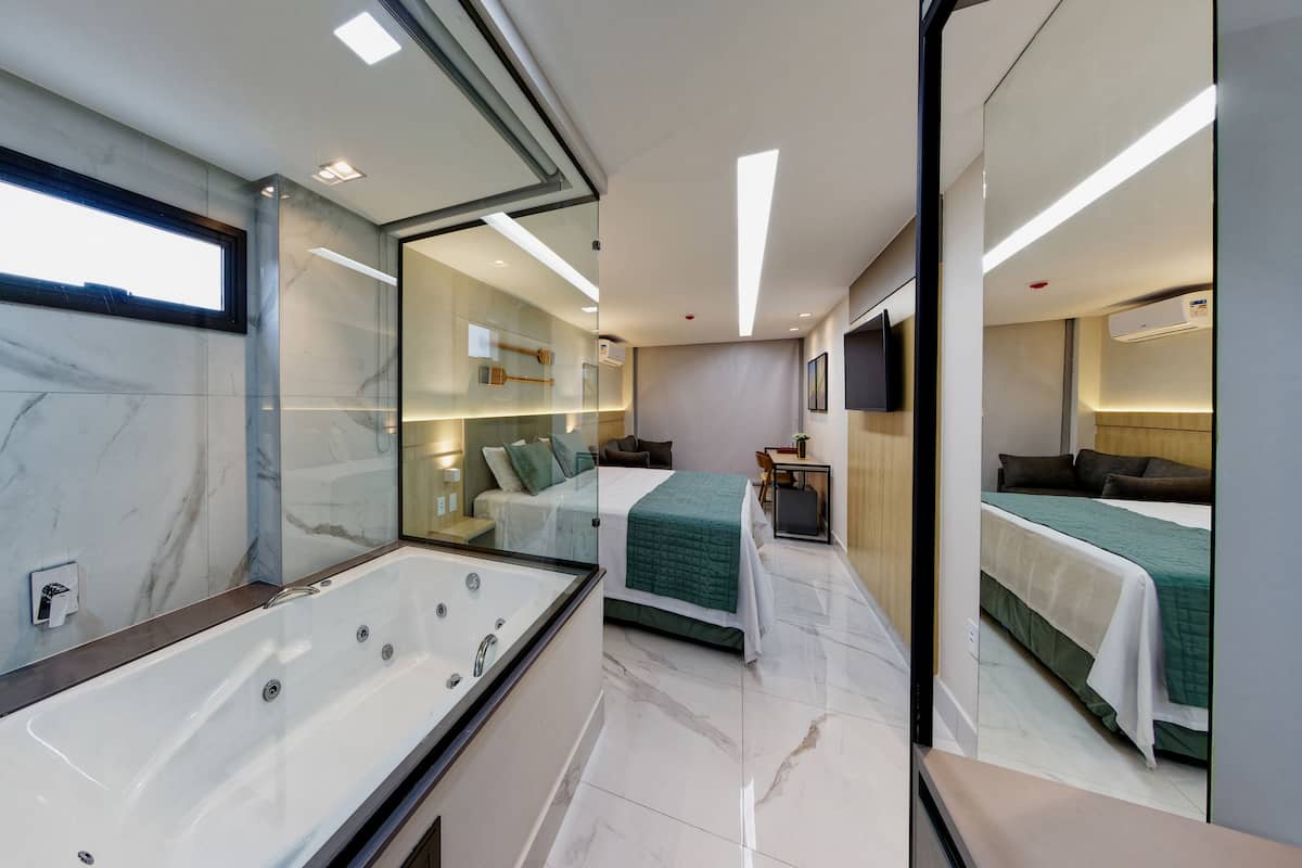 A modern hotel room with a large mirror, a bathtub, a bed with green bedding, and a small desk area.