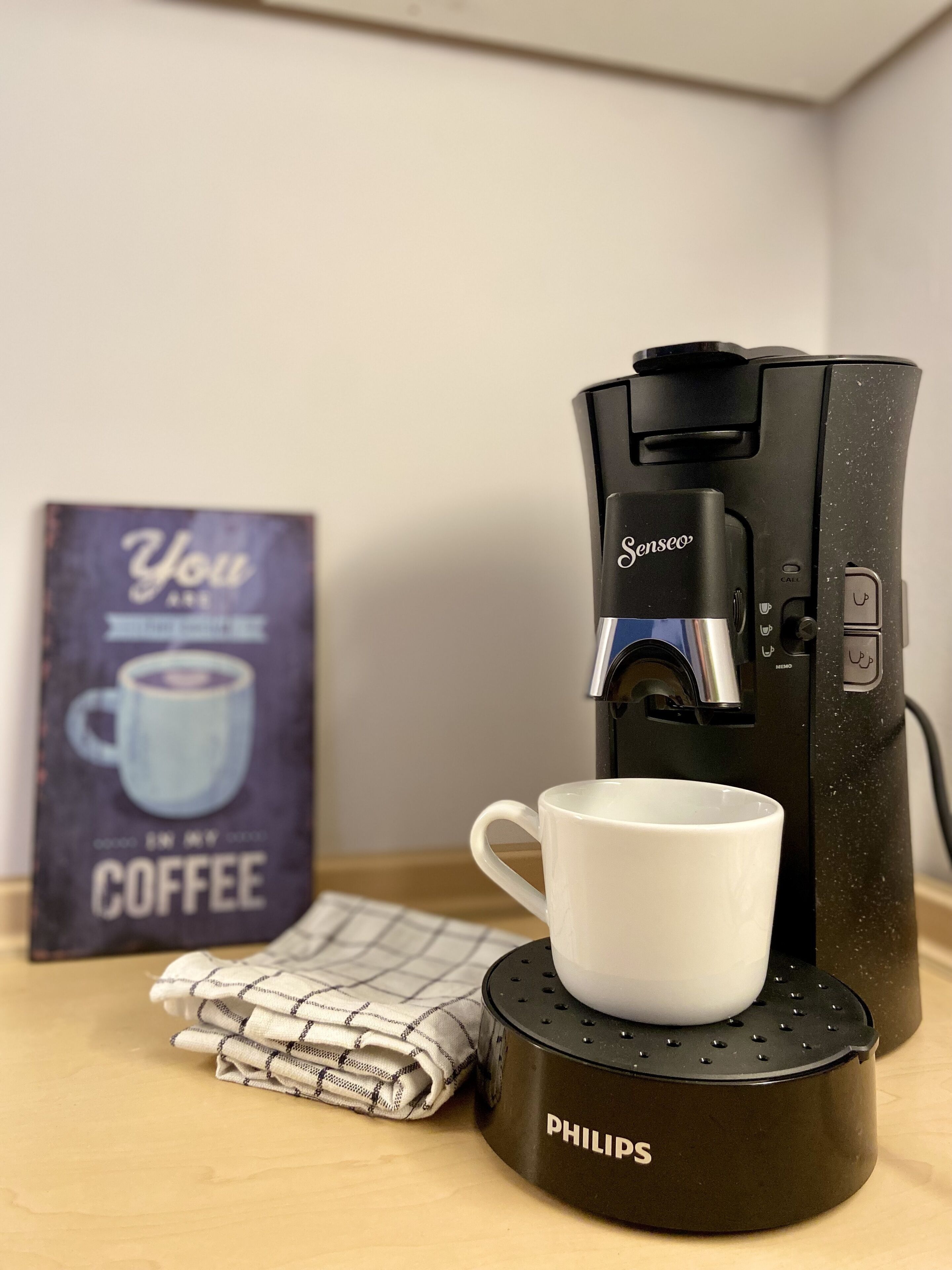 Una macchina da caffè su una base nera, con una tazza bianca sopra. Si vedono un sottopiatti e un cartello con una tazza di caffè.