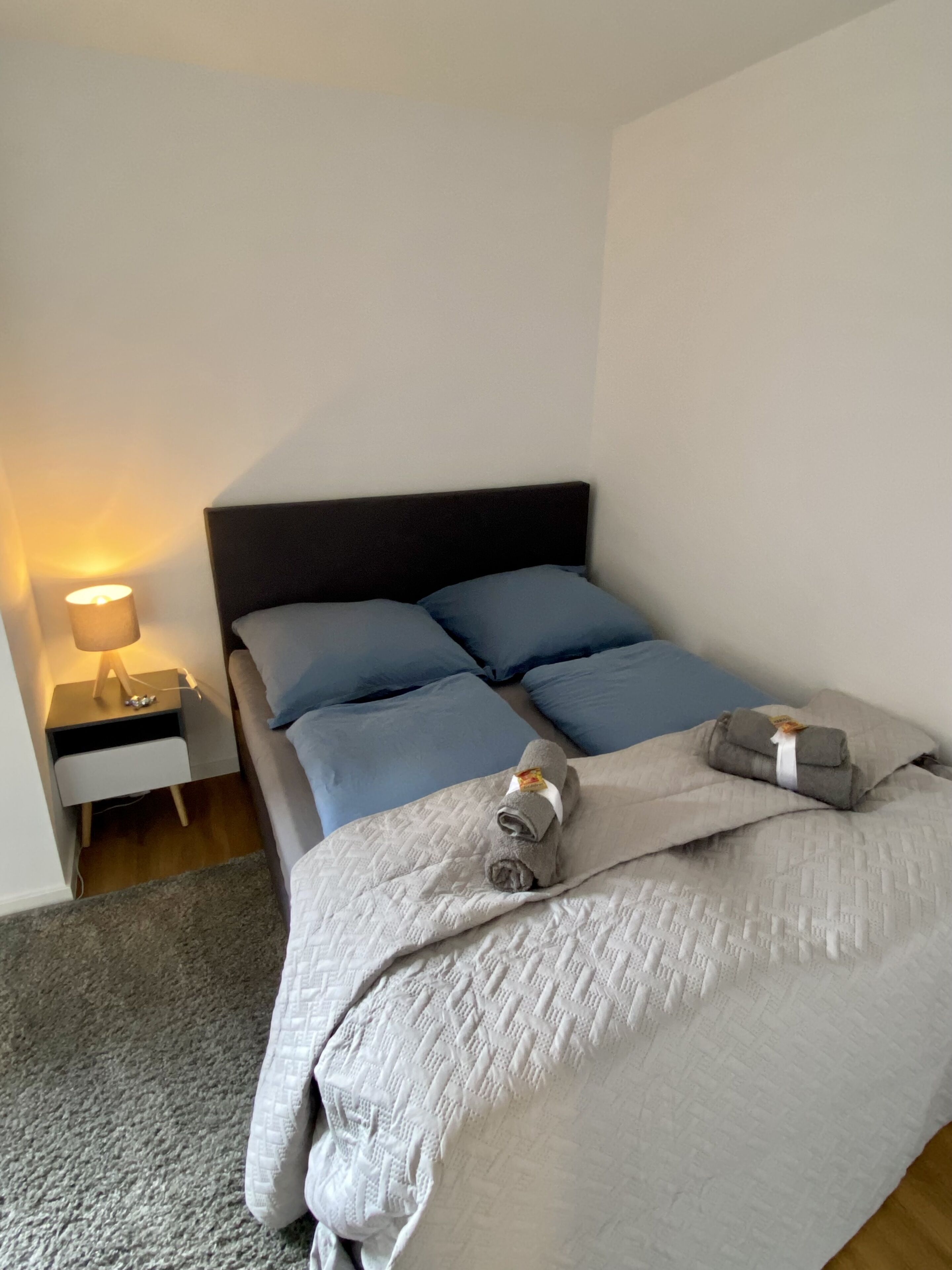 Un lit bien fait, avec une couverture grise et des oreillers, une lampe de chevet et une petite table de nuit en bois sur laquelle se trouve une lampe.
