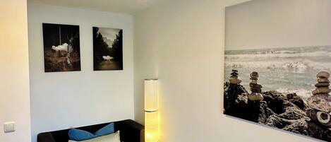 A bedroom with a bed, a bedside table, a wall-mounted lamp, and two framed pictures.