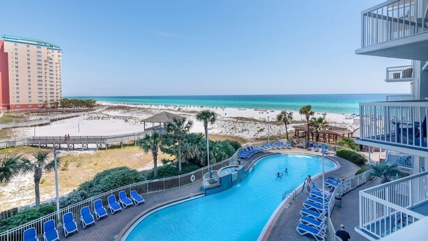 A pool area with a view of the beach and ocean, surrounded by lounge chairs and a building with balconies. - Pelican Beach 0413 2 Bedroom Condo by Pelican Beach Management (Destin)