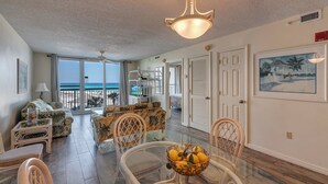 A spacious living room with a large glass door leading to a balcony overlooking the sea, furnished with a sofa, chairs, and a dining table with a fruit bowl. - Pelican Beach 0413 2 Bedroom Condo by Pelican Beach Management (Destin)