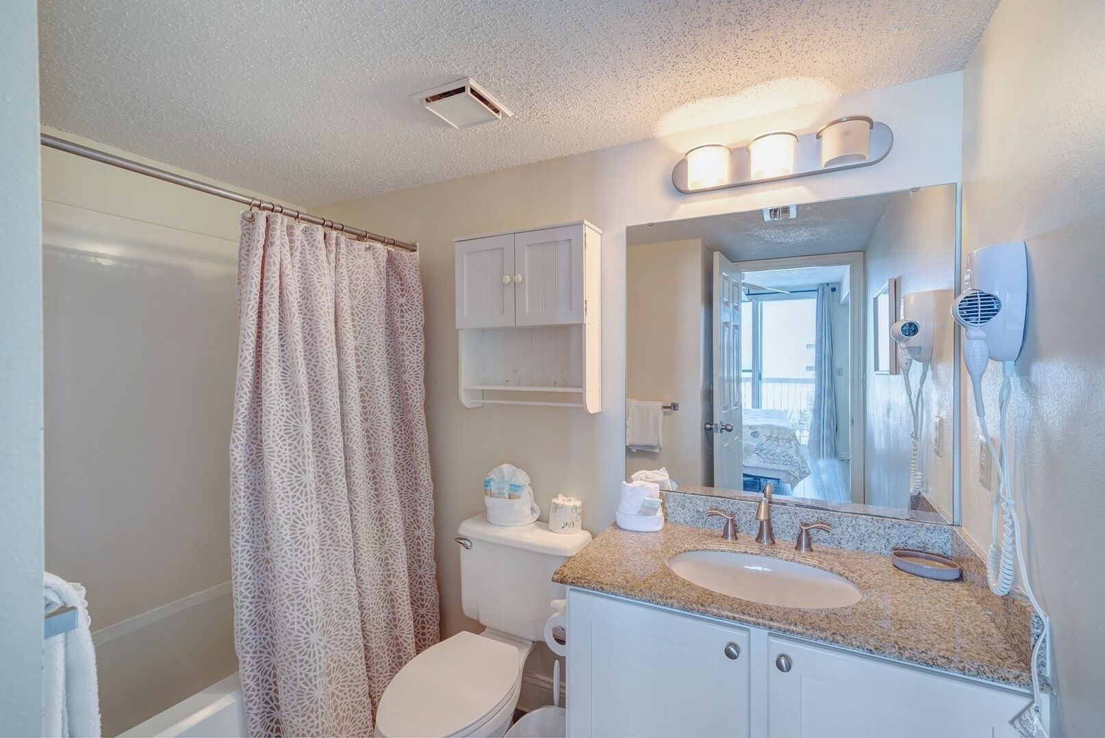 A bathroom with a shower curtain, toilet, sink, and a hairdryer mounted on the wall.