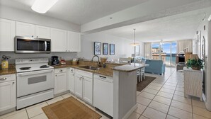 A modern kitchen with white cabinets, a built-in microwave, and a stove. There is a dining area with a blue sofa and a view of the sea. - Pelican Beach 1816 2 Bedroom Condo by Pelican Beach Management (Destin)