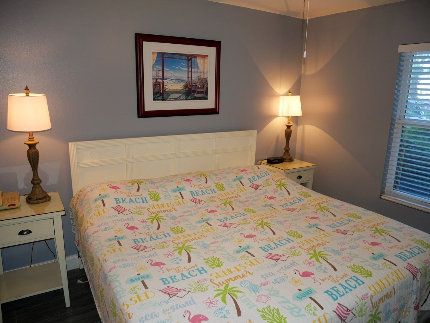 A bedroom with a bed, two bedside tables, a nightstand, a framed picture on the wall, and a window with blinds.