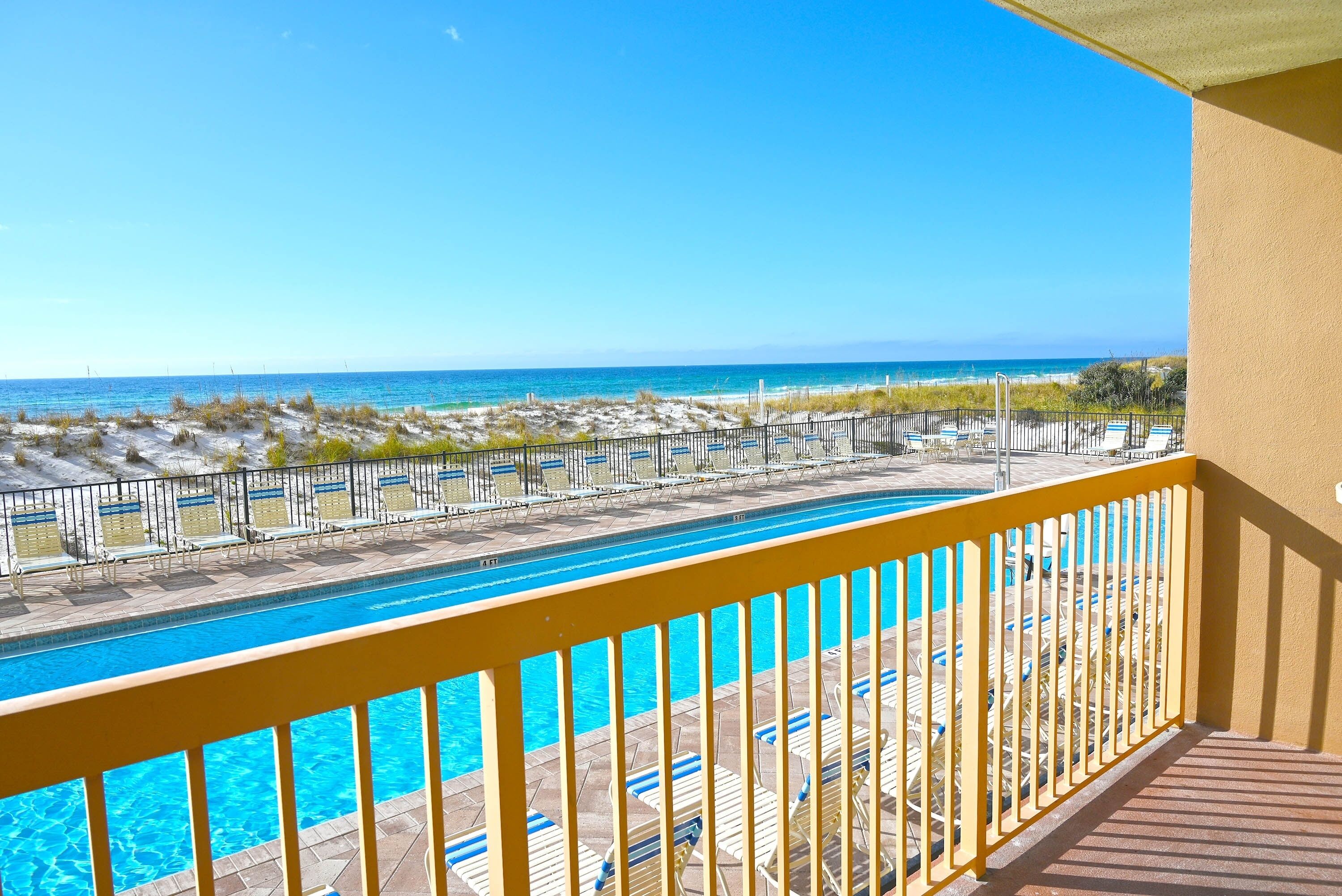 A pool with clear blue water, surrounded by lounge chairs and umbrellas, overlooking a beach with a fence.