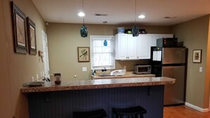 A kitchen with a central island, bar stools, a microwave, a refrigerator, and a window with blinds. - Leafy, Suburban Abode (Greensboro)
