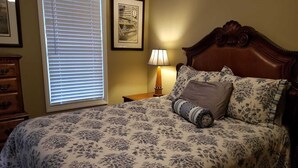A bedroom with a bed, a nightstand, a lamp, a dresser, and a window with blinds. - Leafy, Suburban Abode (Greensboro)