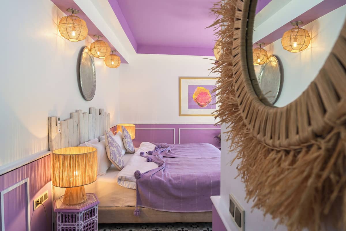 A bedroom with a purple and white color scheme, a bed with a purple blanket, a wall-mounted mirror, and decorative wall lights.