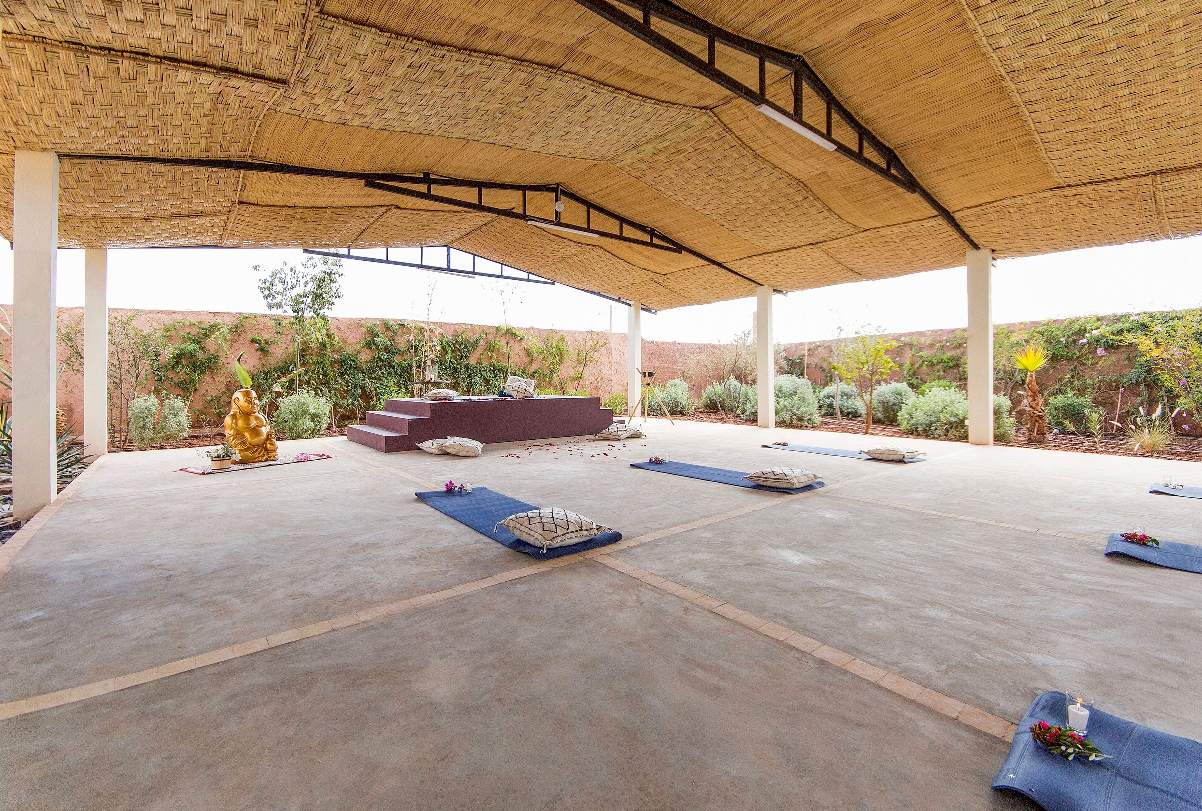 An outdoor covered area with a concrete floor, a golden statue, and a seating arrangement with cushions and mats.