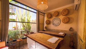 A spa room with a massage table, a window with a view of cacti, and decorative woven wall art. - Maison Daouia (Marrakech)