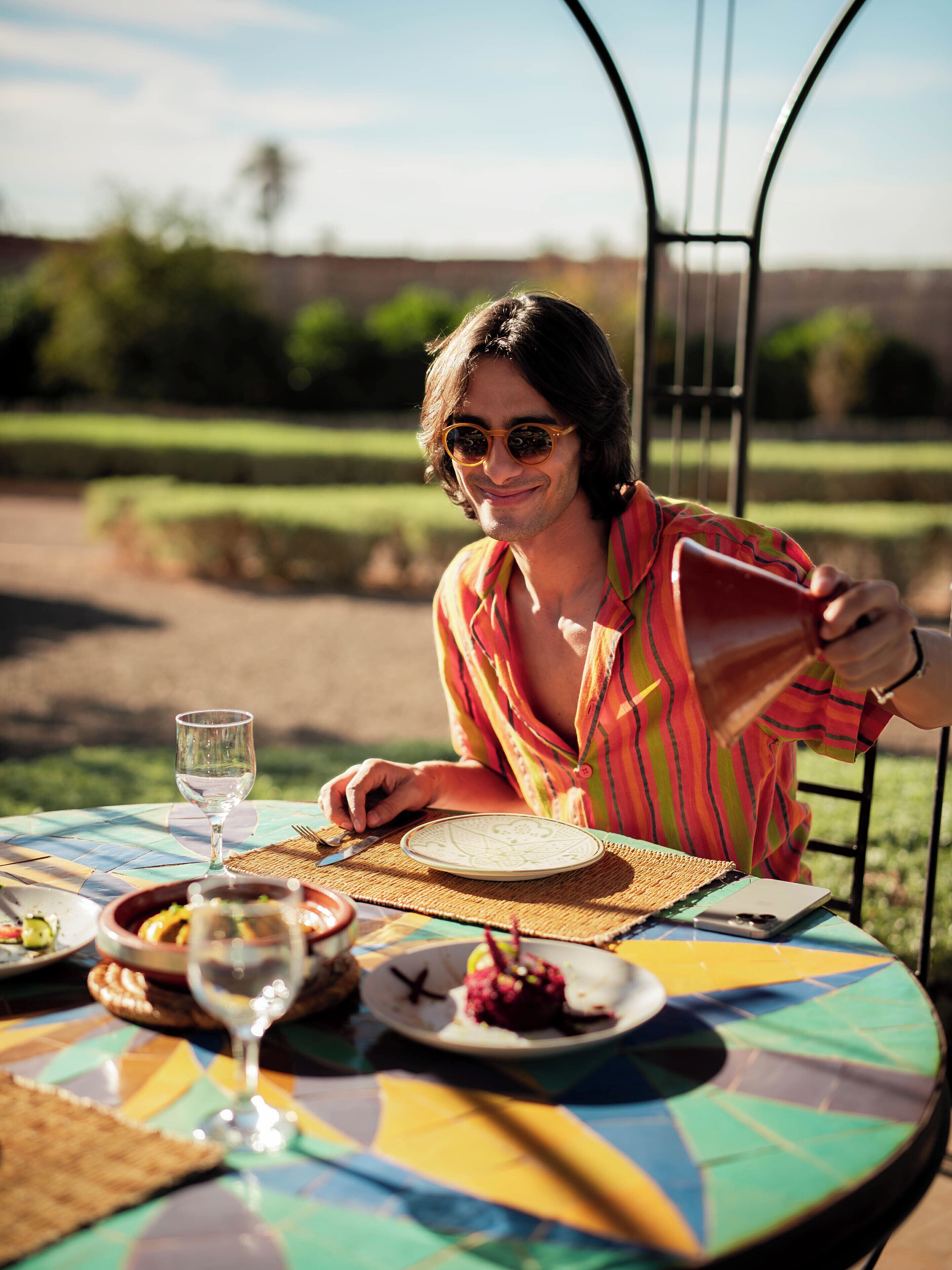 A person seated at an outdoor table with a colorful tablecloth, a glass of water, and plates with food.