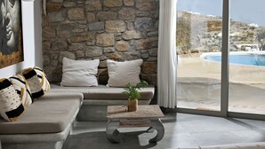 A modern living room with a stone wall, a beige sofa, two white pillows, a striped throw, and a view of a pool and hillside. - Paraga Scorpios Villa 2 7mins Walk to Beach by Calypso Sunset Villas (Mykonos)