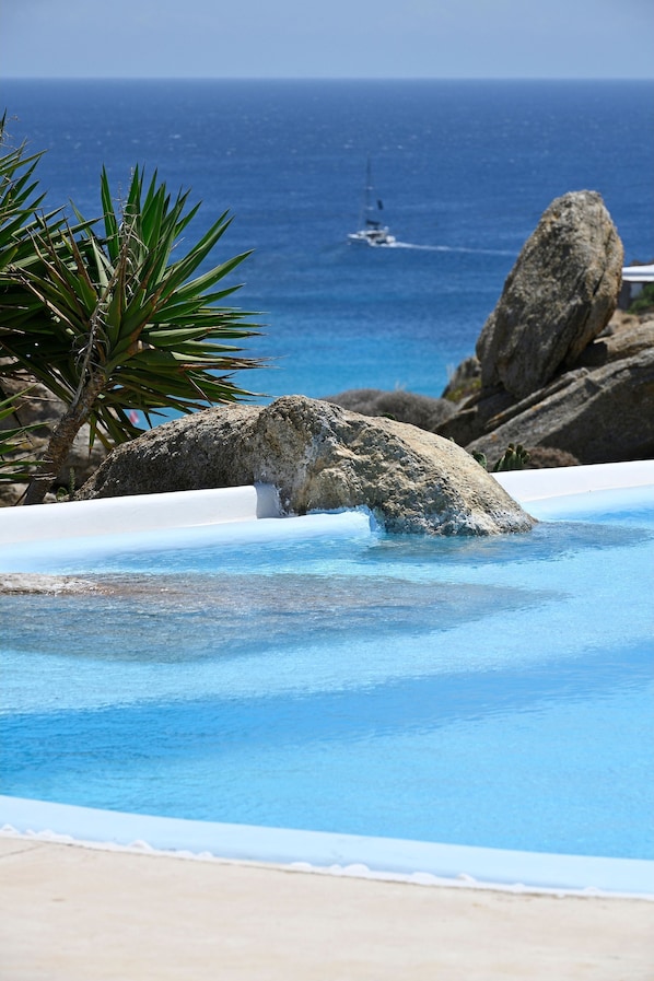 Une piscine bordée de rochers, avec une eau d’un bleu cristallin, et une vue sur l’océan où l’on aperçoit un voilier au loin.