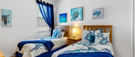 Un dormitorio con dos camas, cada una con ropa de cama azul y blanca, una mesita de noche con una lámpara y tres obras de arte colgadas en la pared que representan tortugas marinas.