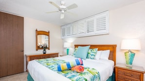 A bedroom with a bed, a ceiling fan, a window with shutters, a nightstand, and a lamp.