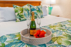 A bed with a tropical leaf-patterned bedspread, a bottle of Veuve Clicquot champagne, two glasses, and a woven basket with a red flower. - Papakea L207 Ocean Front 1 Bedroom 1 Condo by RedAwning (Lahaina)