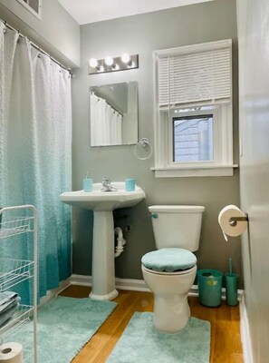 Une salle de bain avec un lavabo sur colonne, des toilettes, une fenêtre avec des stores et un rideau de douche.