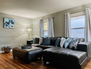 Un salon moderne avec un canapé d’angle, des poufs, une lampe sur pied et un tableau encadré au mur.