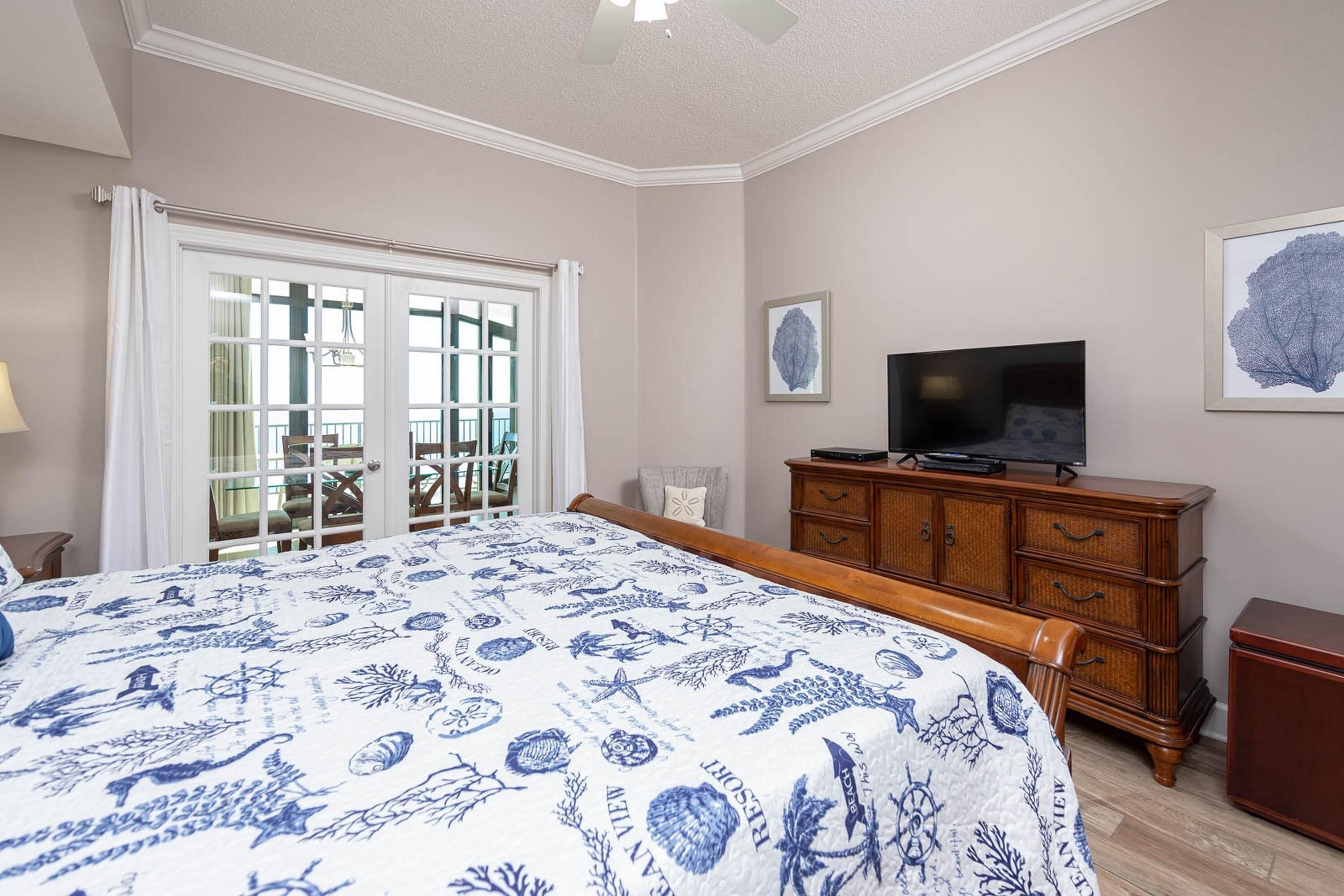 A bedroom with a large bed, a wooden dresser, a television, and a balcony with a view.