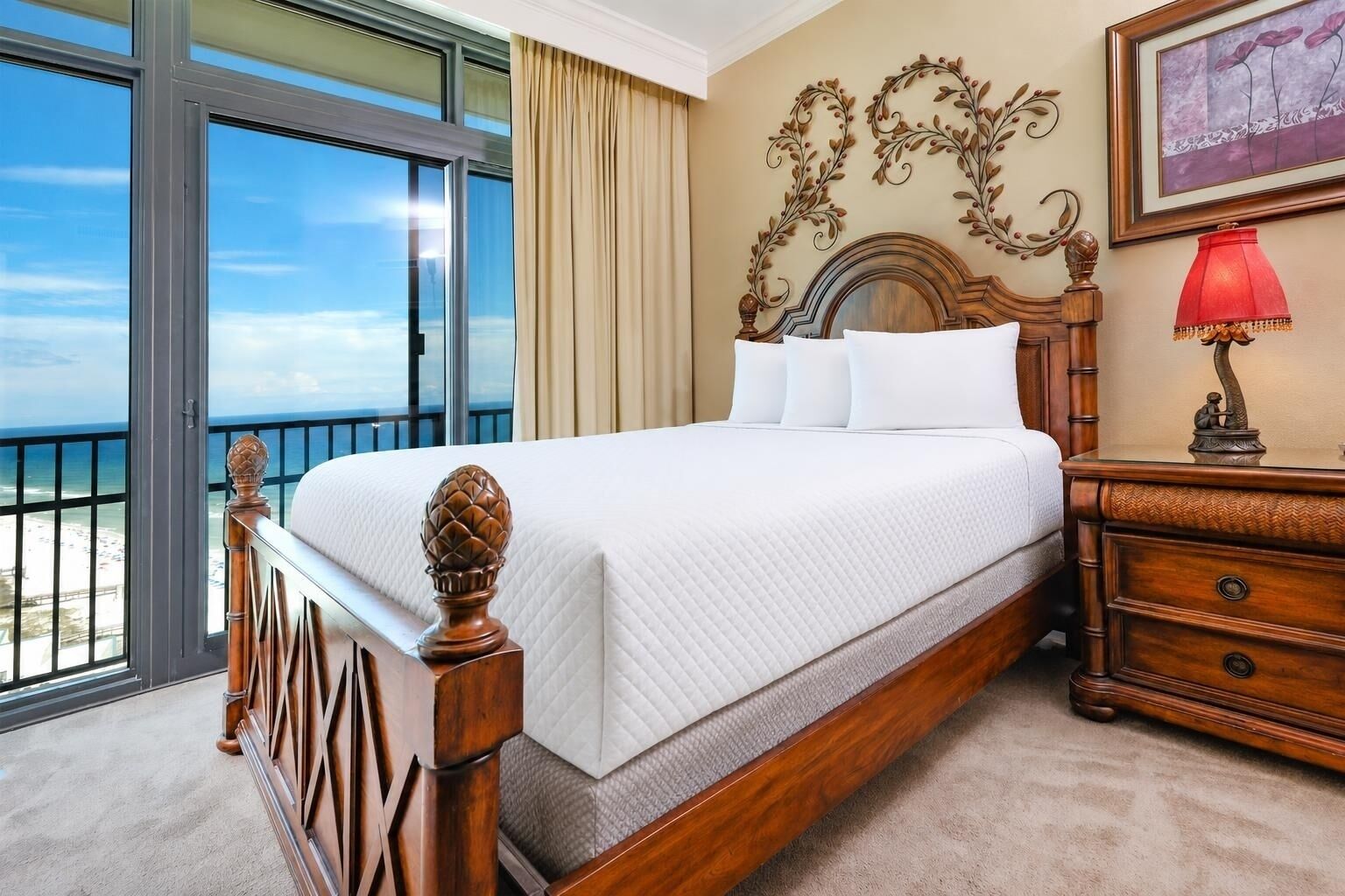 A hotel room with a large bed, a nightstand, a red lamp, and a view of the ocean through a balcony.