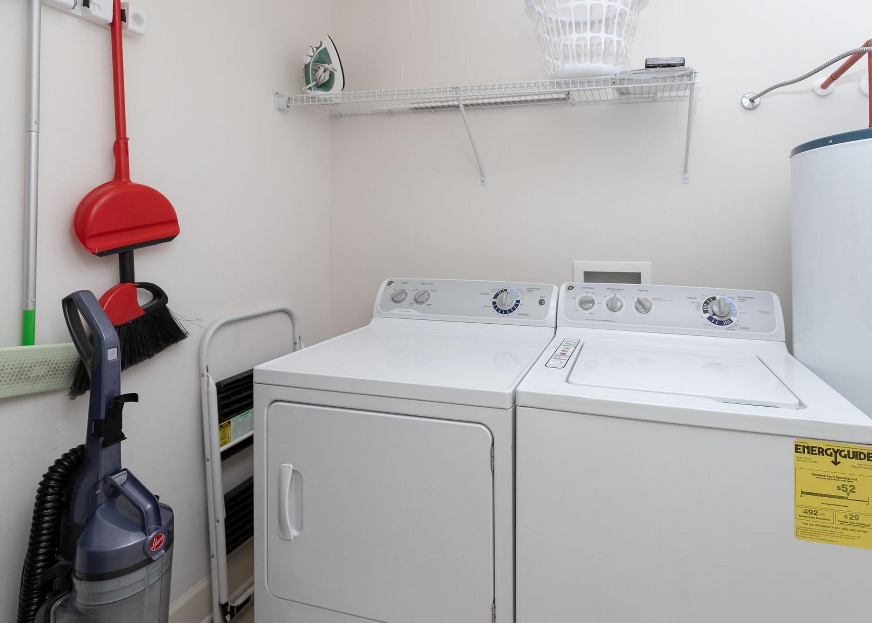 Une buanderie équipée de deux machines à laver blanches, d’une serpillière rouge, d’un aspirateur et d’une petite étagère sur laquelle se trouve un panier.