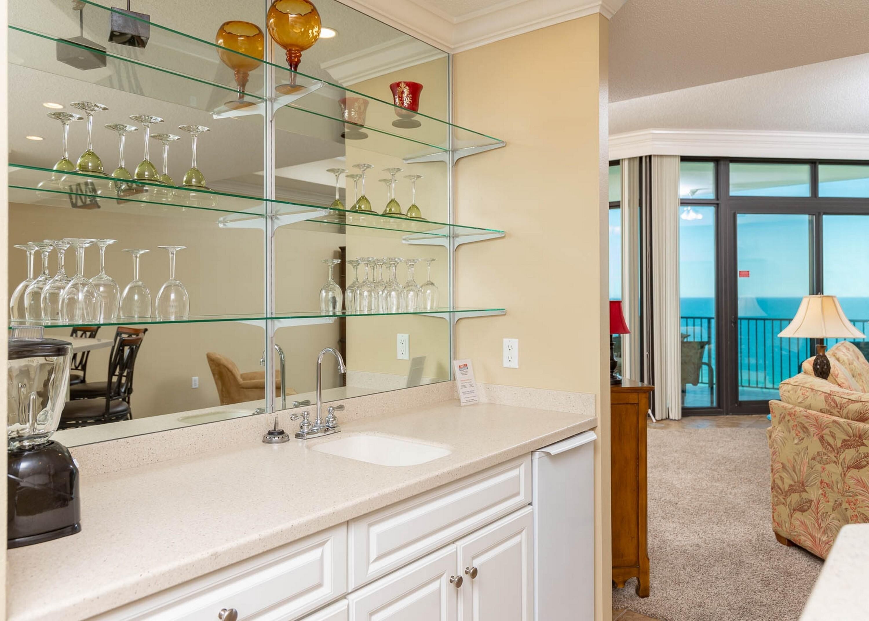 A hotel room with a large mirror, a sink, and a glass shelf with wine glasses. There is a balcony with a view of the ocean.