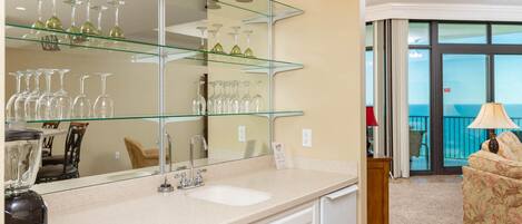 A hotel room with a large mirror, a sink, and a glass shelf with wine glasses. There is a balcony with a view of the ocean.