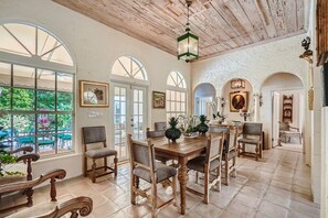A dining area with a wooden table and chairs, arched windows, and a view of greenery outside.