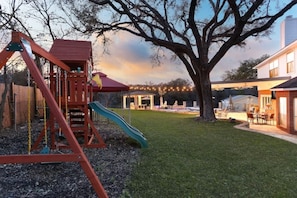 A playground with a swing set and slide, a large tree, and a house with outdoor seating.
