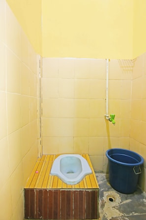 Un baño pequeño y cerrado con un asiento de madera para el inodoro, un cubo azul y un cabezal de ducha.