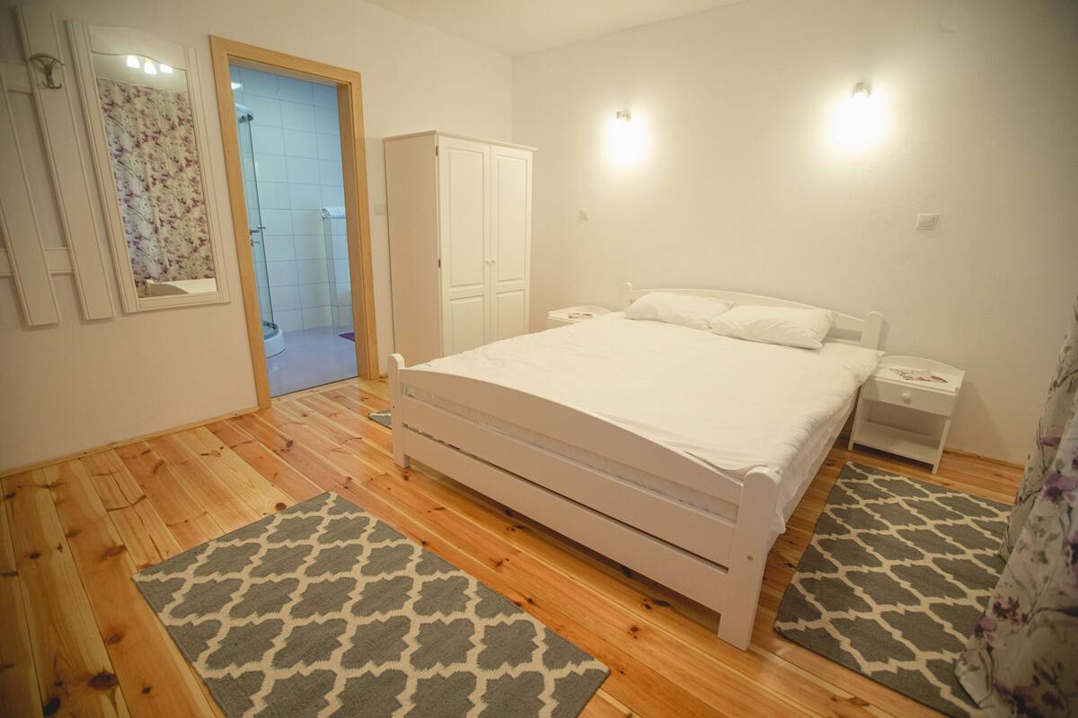 A bedroom with a wooden floor, a white bed, a nightstand, a wardrobe, and a bathroom visible through an open door.