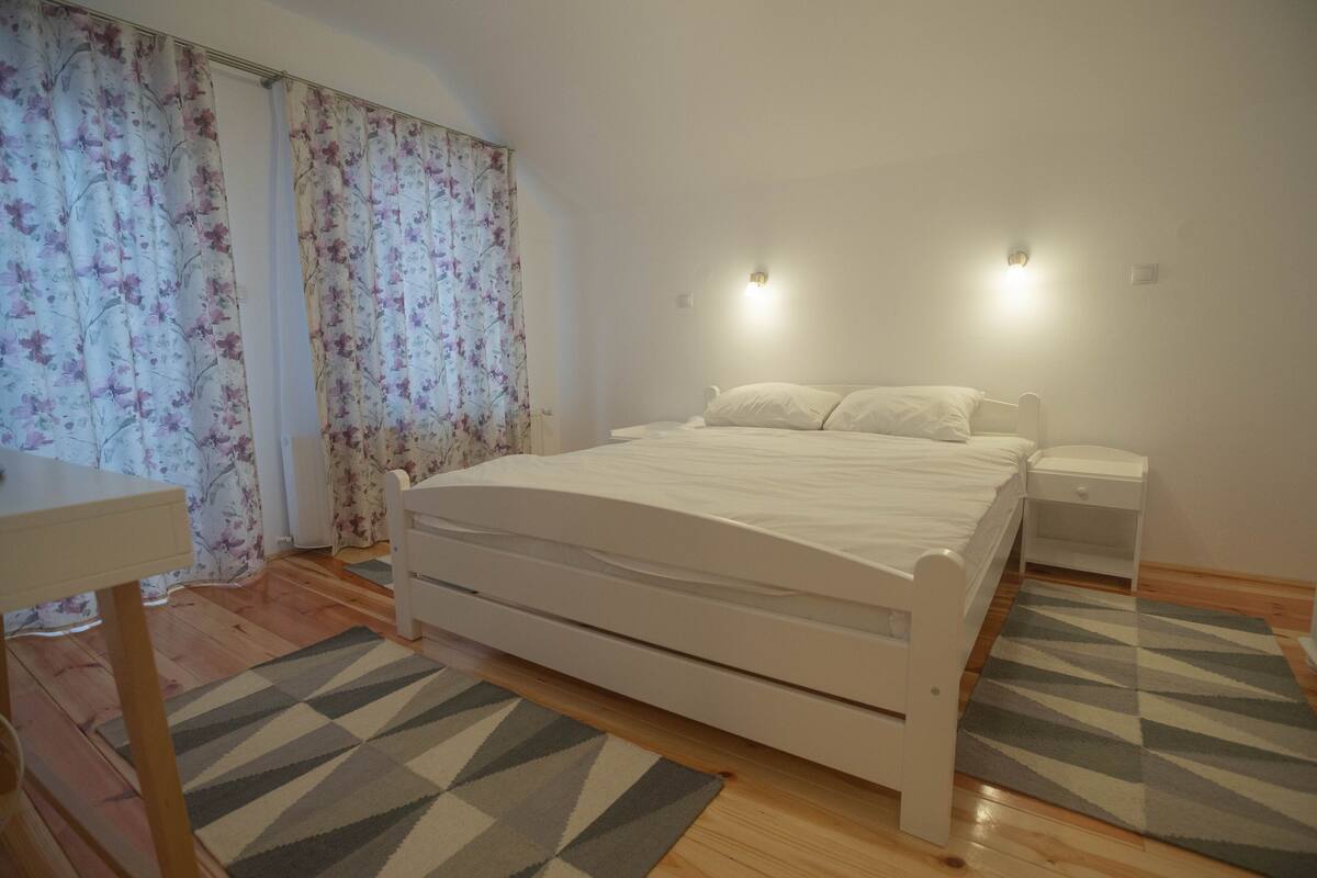 A bedroom with a bed, bedside tables, a desk, and curtains.