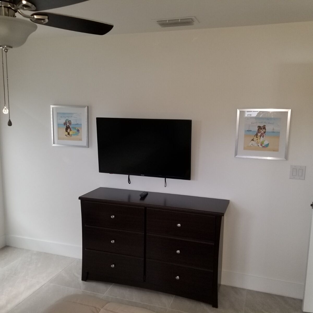 Una habitación con una televisión de pantalla plana montada en la pared, un aparador de madera oscura con cajones y dos cuadros enmarcados.