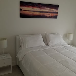 A bedroom with a bed, white bedding, a nightstand with a lamp, and a painting of a sunset on the wall.