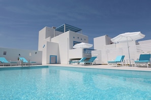 En pool med solstolar och parasoller, en vit byggnad med geometriskt mönster och en klarblå himmel.