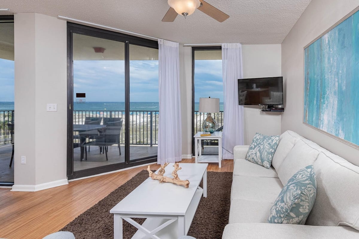 A living room with a white sofa, a coffee table, and a TV on the wall. There is a balcony with a view of the beach and ocean.