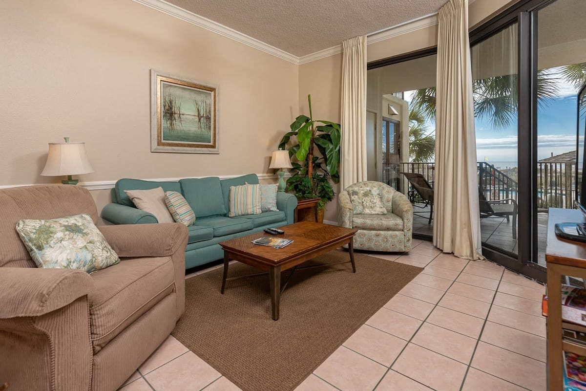 A living room with a sofa, armchair, coffee table, and a large window with a view of palm trees.